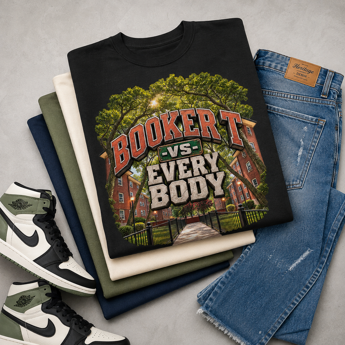 A folded black graphic T-shirt is arranged on top of stacked neutral-colored T-shirts, blue jeans, and sneakers on a light gray surface. The shirt features a large graphic of a red brick building under green trees with the text 'BOOKER T -VS- EVERY BODY'.