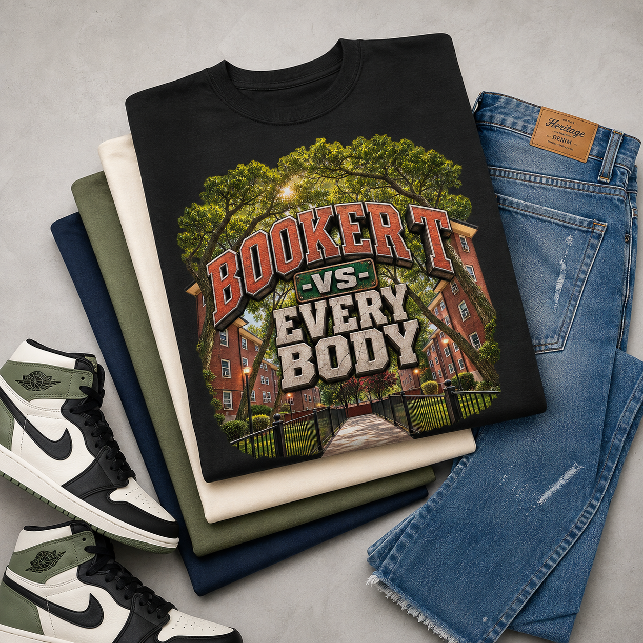 A folded black graphic T-shirt is arranged on top of stacked neutral-colored T-shirts, blue jeans, and sneakers on a light gray surface. The shirt features a large graphic of a red brick building under green trees with the text 'BOOKER T -VS- EVERY BODY'.